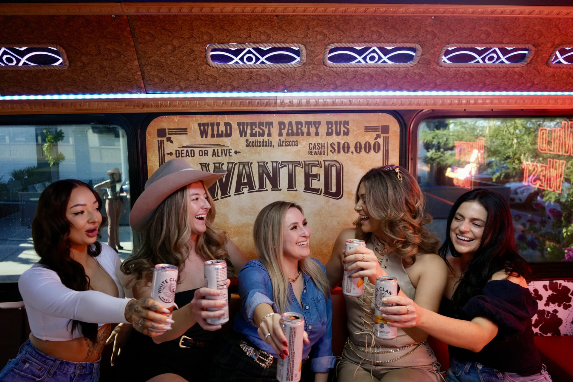 Five guests laughing and toasting drinks in front of the Wild West Party Bus wanted poster